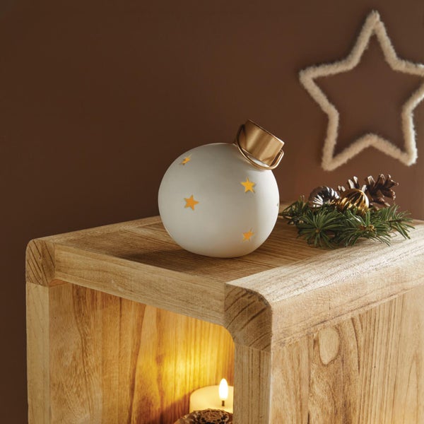 Weiße weihnachtliche Dekokugel aus Keramik mit Sternausstanzungen und goldfarbenem Aufhänger auf einem hölzernen Beistelltisch.