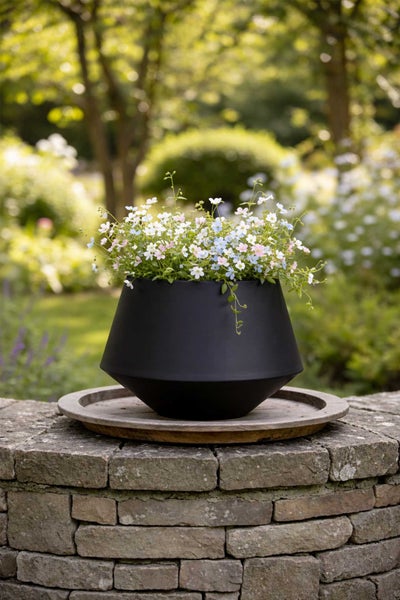 Schwarzer matter Pflanztopf mit kleinen Blumen auf einer Steinmauer in einem hellen Garten.