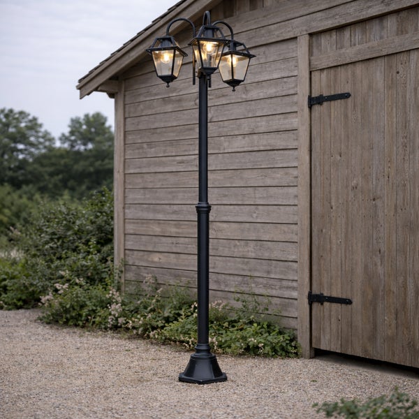 Dreiflammige Außenleuchte in Schwarz auf einem Kiesweg vor einer Holzhütte.
