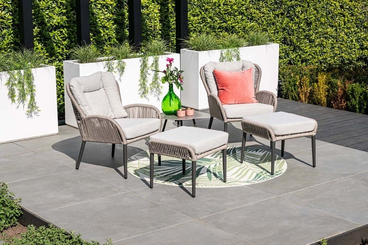 Garten-Lounge-Set aus zwei Sesseln mit Polstern, zwei Fußhockern und einem Beistelltisch auf einer Terrasse vor Pflanzgefäßen und einer Hecke.