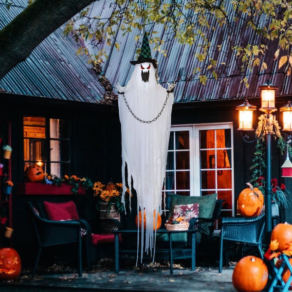 Dekorative Halloween-Szene mit einem hängenden Geist mit Hexenhut und Ketten vor einem Haus mit Kürbissen und herbstlicher Dekoration.