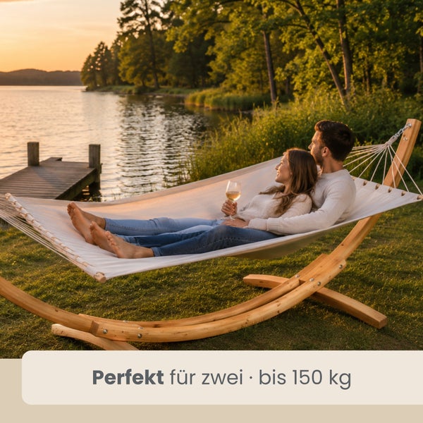 Paar entspannt in weißer Hängematte mit Holzgestell an einem See bei Sonnenuntergang. Text: Perfekt für zwei, bis 150 Kilogramm.