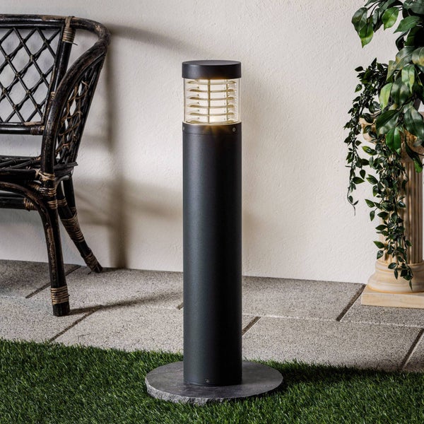 Moderne anthrazitfarbene Pollerleuchte für den Außenbereich mit beleuchtetem Glaseinsatz und Lamellen auf einer Steinterrasse im Garten.