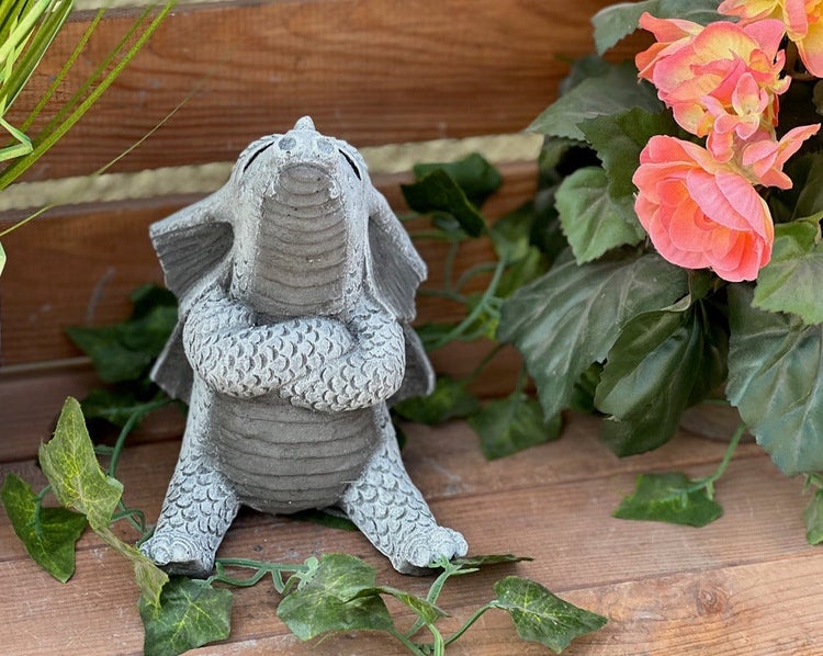 Kleine Gartenfigur Drache in Steinoptik mit verschränkten Armen und nach oben gerichtetem Blick, umgeben von Pflanzen auf einer Holzoberfläche.