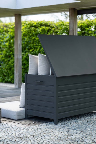 Anthrazitfarbene Gartenbox aus Metall mit geöffnetem Deckel, gefüllt mit hellgrauen Kissen auf einer Terrasse vor einer Hecke.