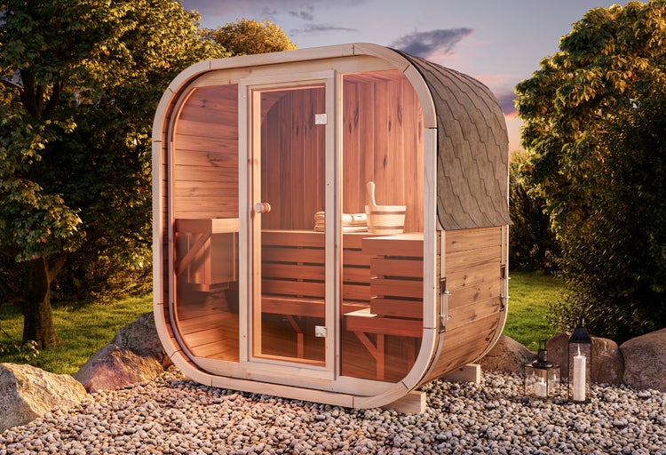 Moderne quadratische Fasssauna aus Holz mit Glasfront, Holzbänken und Schindeldach in einer hellen Gartenumgebung auf Kies.