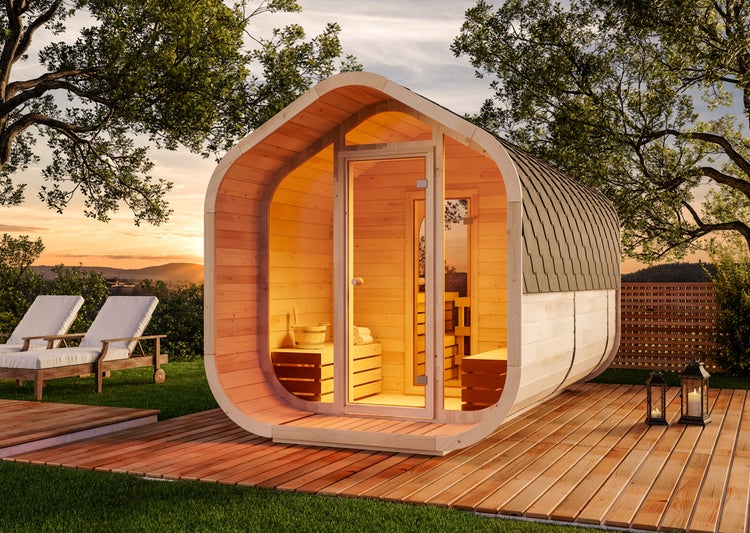 Moderne Fasssauna aus hellem Holz mit Glasfront auf einer Holzterrasse im Garten bei Sonnenuntergang.